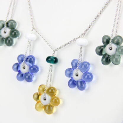 Collier chaine argent 925 avec 5 perles en forme de fleur marguerite de couleur variées, jaune pour celle du milieu puis sur les côtés bleu et grise. Perle fleur transparente. Les perles en verre sont pendantes de part et d'autre de la chaine, tombantes sur le buste. -Atelier horizon verre