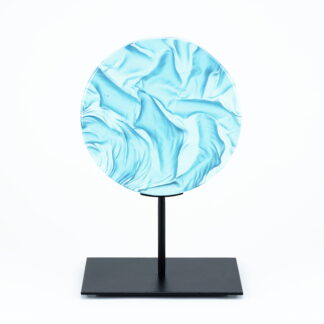 Pièce d'art décoratif de forme circulaire en cristal nommé "Magma" de taille moyenne (18 cm de diamètre). Objet avec du relief, texturés, avec des effets de drapé. Pièce en cristal de couleur bleu ciel sur support en métal thermolaqué noir. Pâte de verre. -Atelier horizon verre