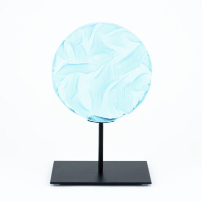 Pièce d'art décoratif de forme circulaire en cristal nommé "Magma" de taille moyenne (18 cm de diamètre). Objet avec du relief, texturés, avec des effets de drapé. Pièce en cristal de couleur bleu ciel sur support en métal thermolaqué noir. Pâte de verre. -Atelier horizon verre