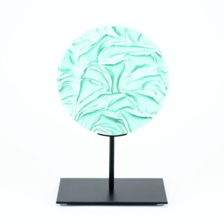 Pièce d'art décoratif de forme circulaire en cristal nommé "Magma" de taille moyenne (18 cm de diamètre). Objet avec du relief, texturés, avec des effets de drapé. Pièce en cristal de couleur vert menthe sur support en métal thermolaqué noir. Pâte de verre. -Atelier horizon verre