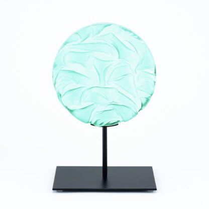 Pièce d'art décoratif de forme circulaire en cristal nommé "Magma" de taille moyenne (18 cm de diamètre). Objet avec du relief, texturés, avec des effets de drapé. Pièce en cristal de couleur vert menthe sur support en métal thermolaqué noir. Pâte de verre. -Atelier horizon verre